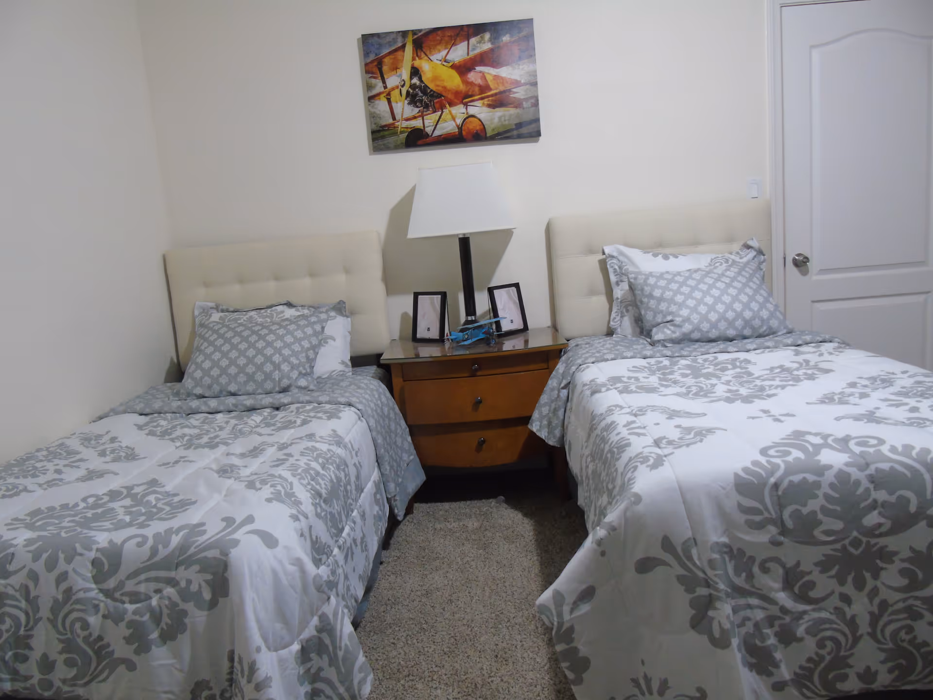 A bedroom with two twin beds featuring white and gray patterned bedding. Between the beds is a wooden nightstand with a white lamp, two small picture frames, and a small blue model airplane. Above the nightstand hangs a colorful painting of a vintage yellow airplane. The room has beige walls and a closed white door on the right side.