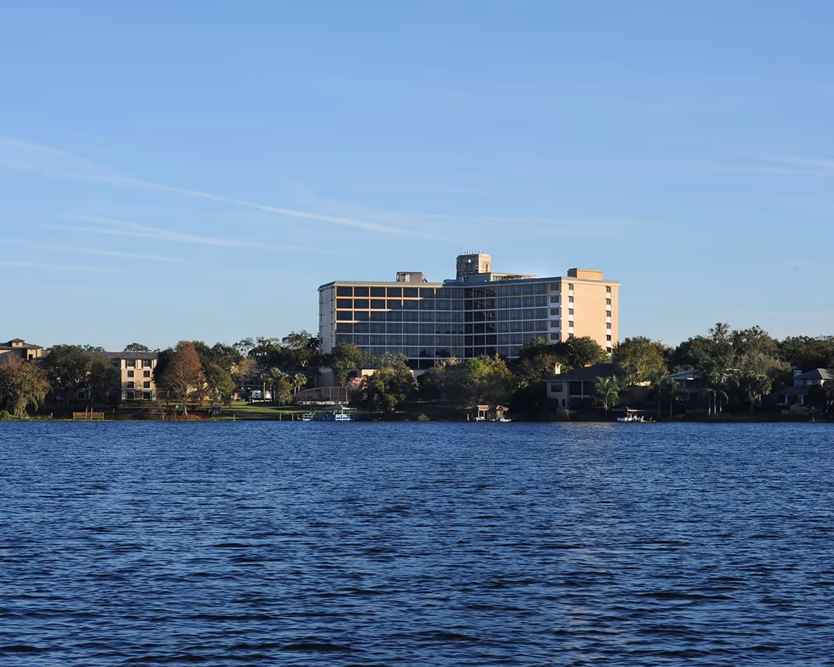 View of a multi-story building situated behind a body of water with trees and smaller buildings surrounding it under a clear blue sky.