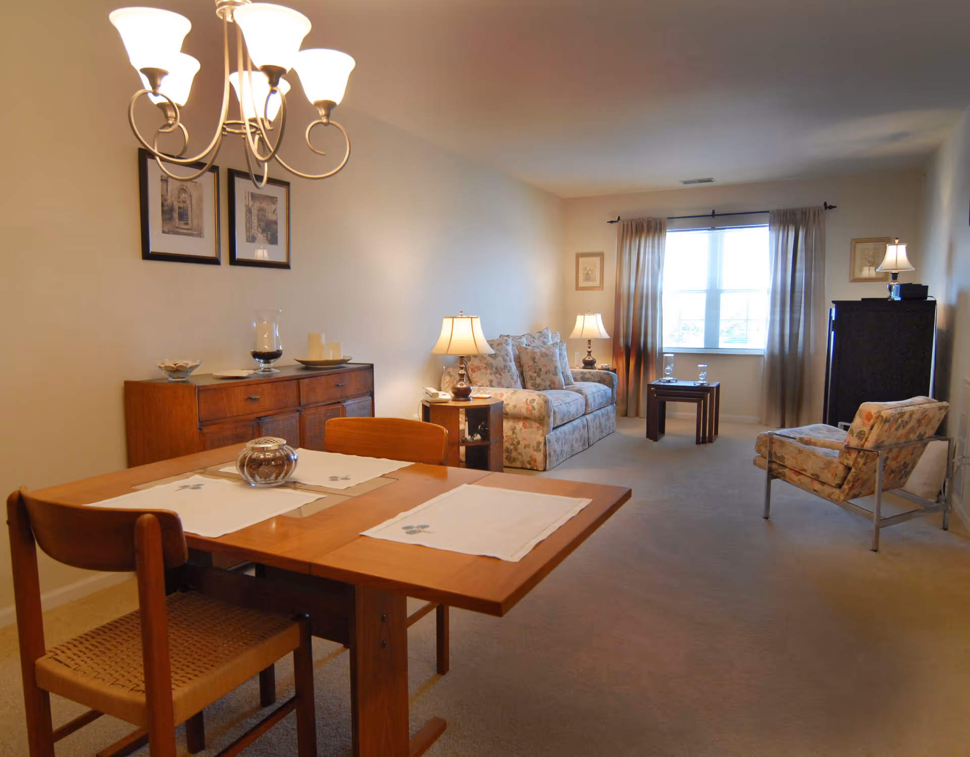 Well-lit combined living and dining area with a wooden dining table in the foreground, floral upholstered sofa and chairs, sideboard, lamps, and a window with curtains.