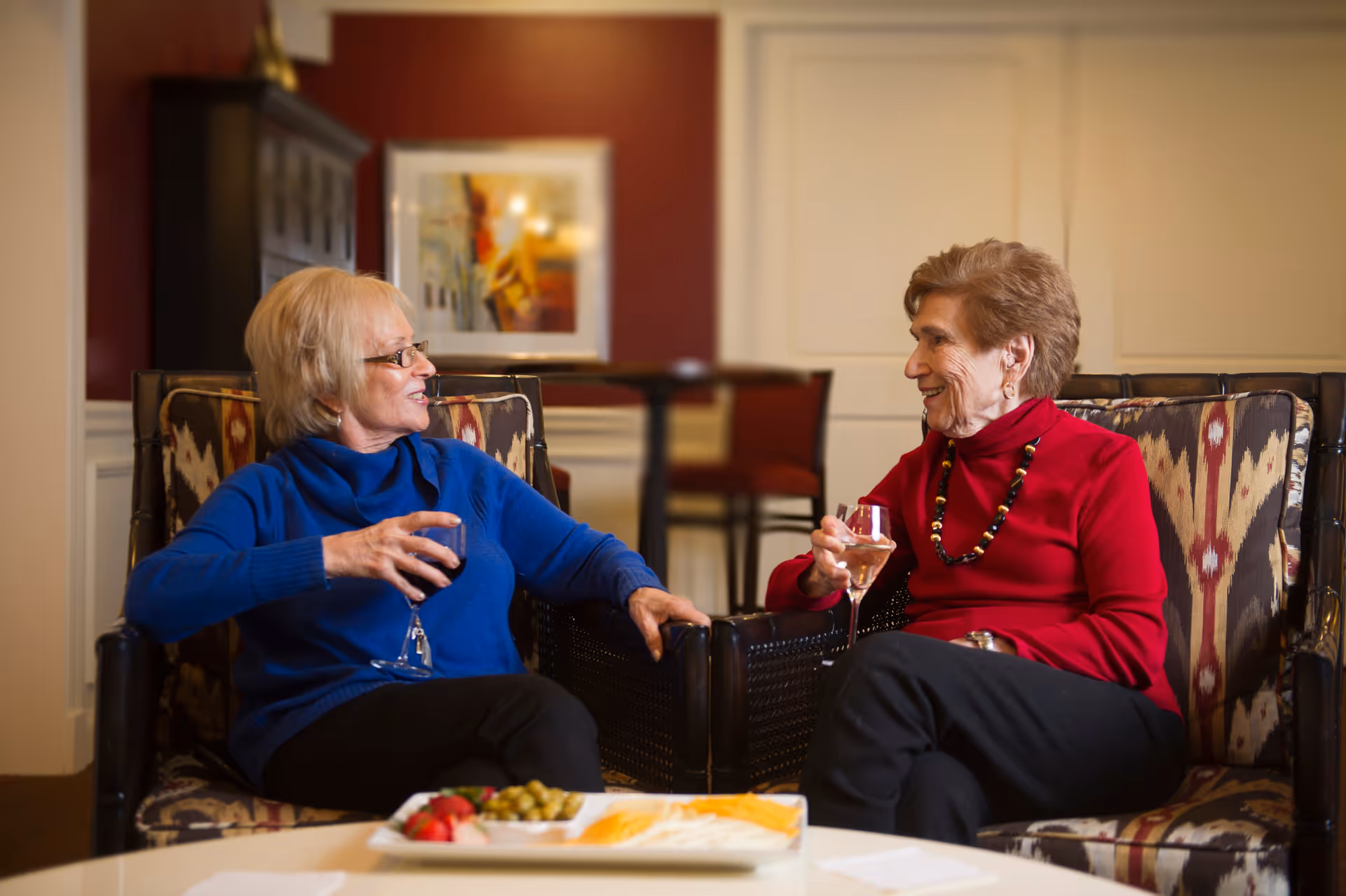 Two elderly women sitting in patterned armchairs in a cozy living room, smiling and holding glasses of wine while chatting. A plate with snacks is on the table in front of them, and there is a framed picture and furniture in the background.