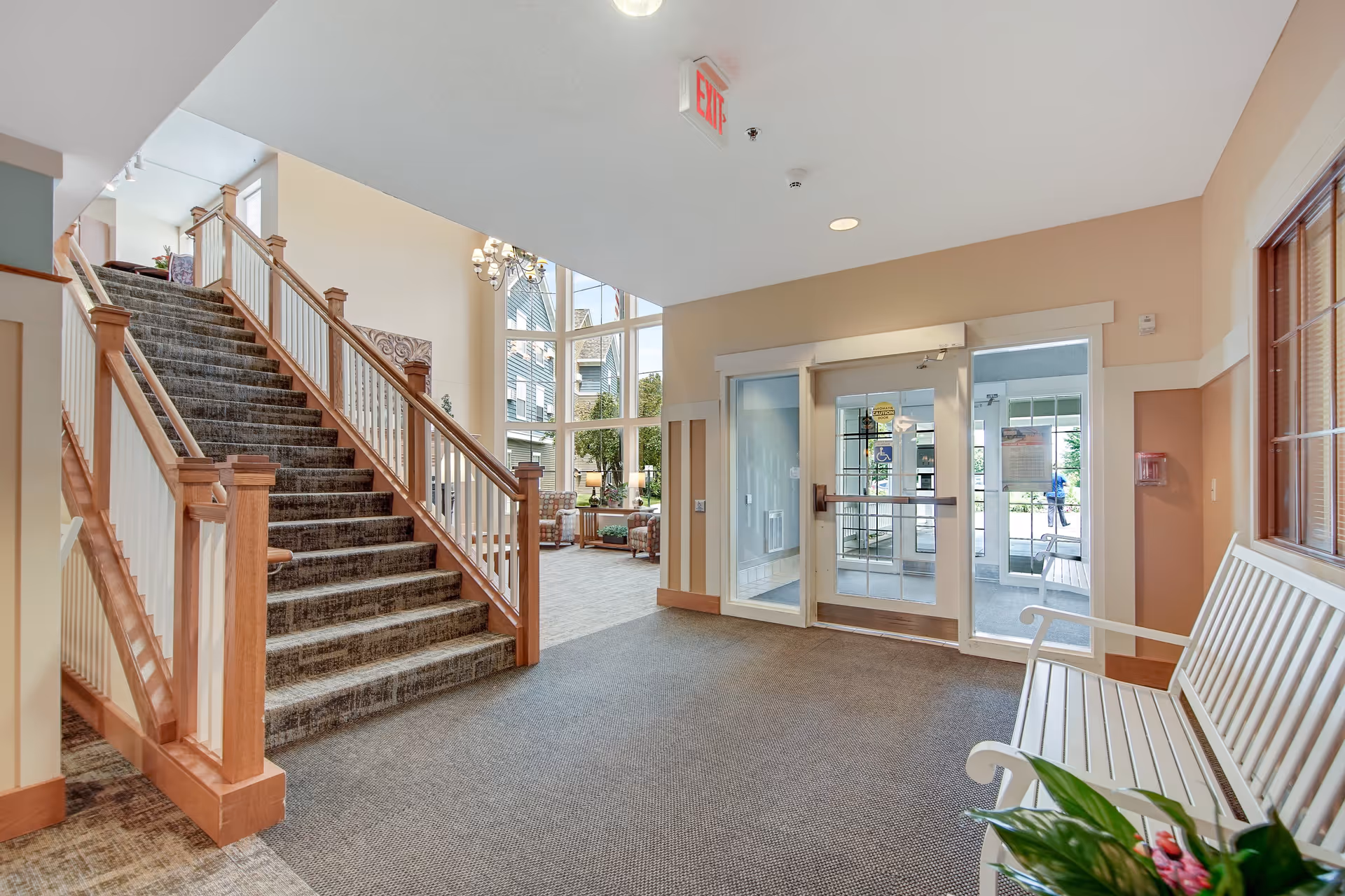 Interior view of an assisted living facility entrance area with a carpeted staircase featuring wooden railings on the left, a glass door exit with an exit sign above it straight ahead, and a white bench with a potted plant on the right. Beyond the staircase, there is a seating area with chairs and large windows letting in natural light.