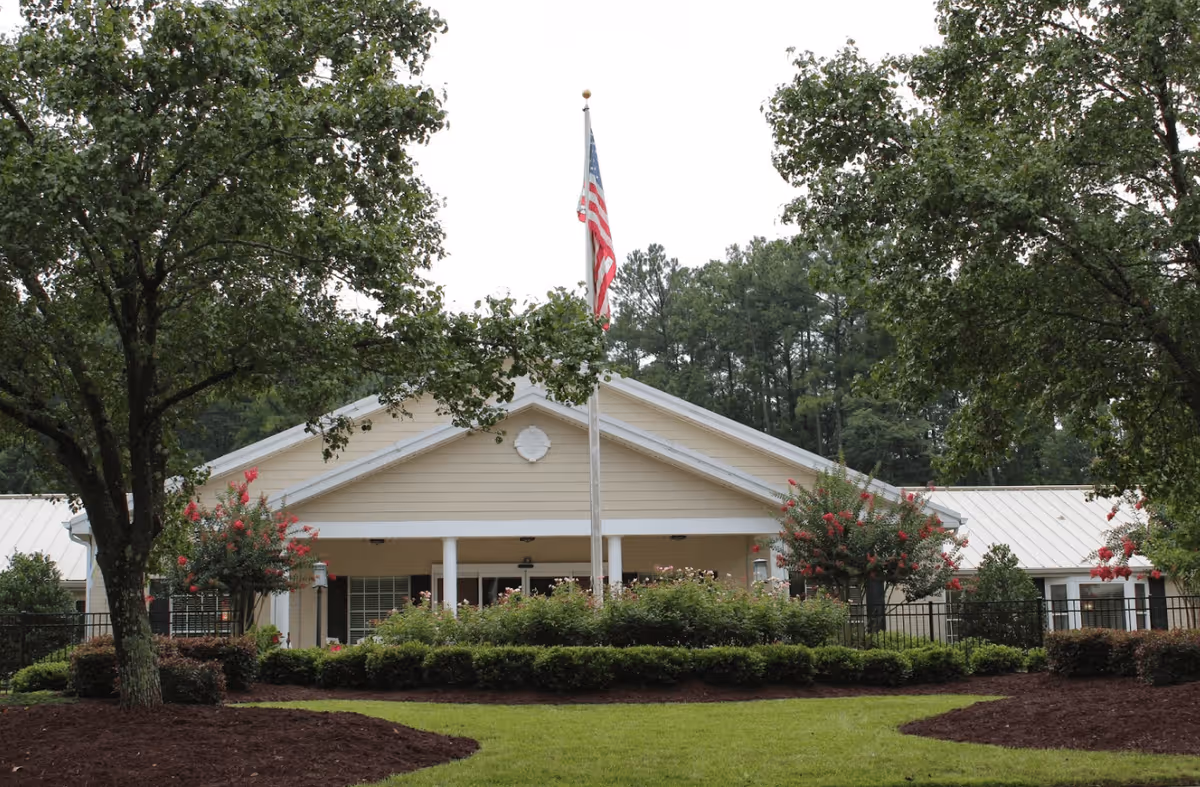 Front exterior view of a single-story building with a beige facade and white roof, surrounded by green trees and shrubs. An American flag is raised on a flagpole in front of the building, with a well-maintained lawn and landscaped garden beds.