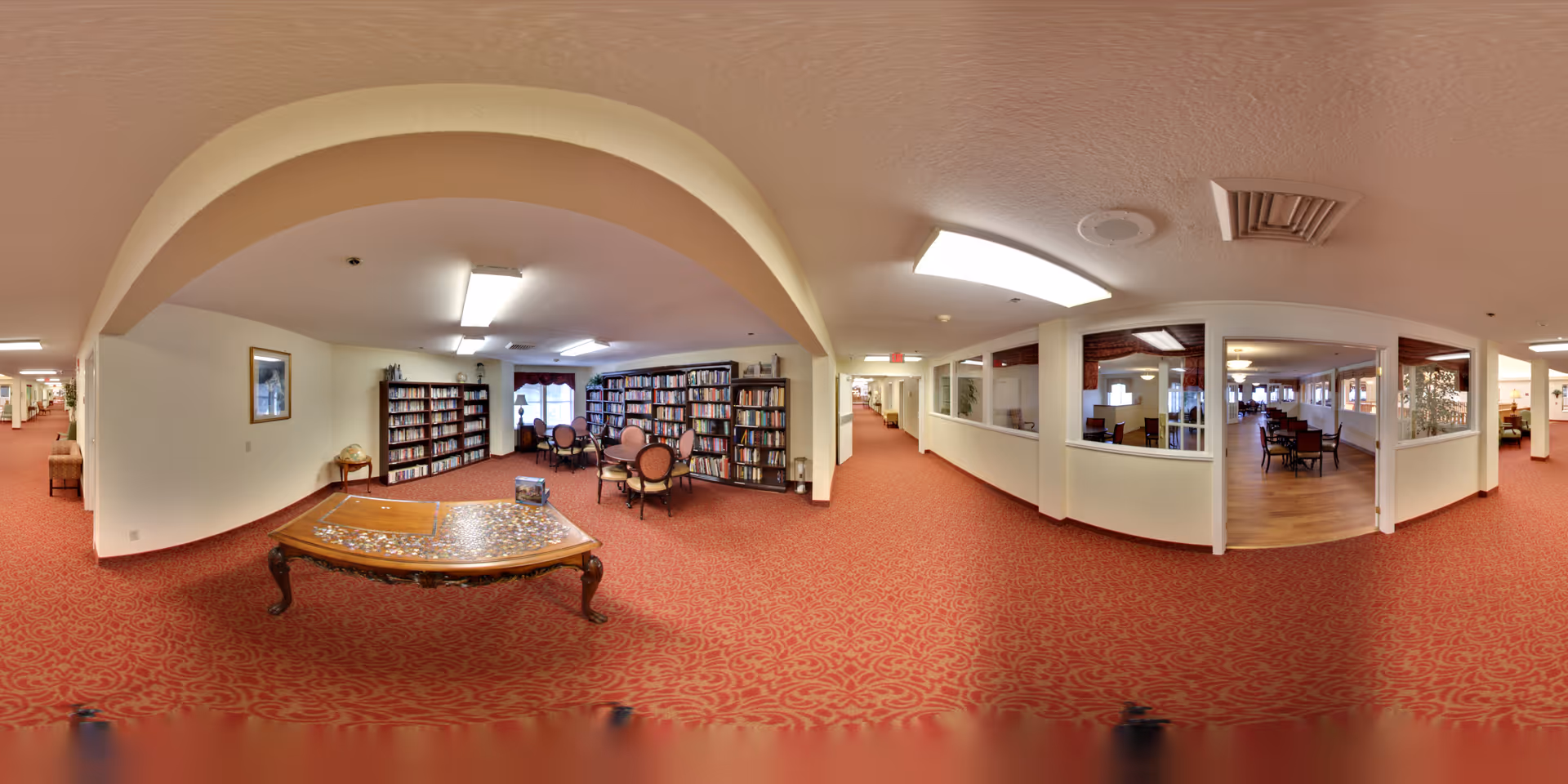A spacious interior hallway with red patterned carpet and white walls. On the left side, there is a cozy library area with bookshelves filled with books and round tables with chairs for reading. On the right side, there are large windows looking into a room with wooden flooring and several tables and chairs arranged for group activities or dining. The ceiling has fluorescent lighting and an archway separates the library area from the hallway.
