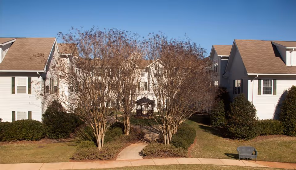Exterior view of a senior living facility with white buildings, brown roofs, and multiple windows. There is a paved walkway leading through landscaped bushes and leafless trees toward a gazebo in the center courtyard. A bench is visible on the grass near the sidewalk in the foreground under a clear blue sky.