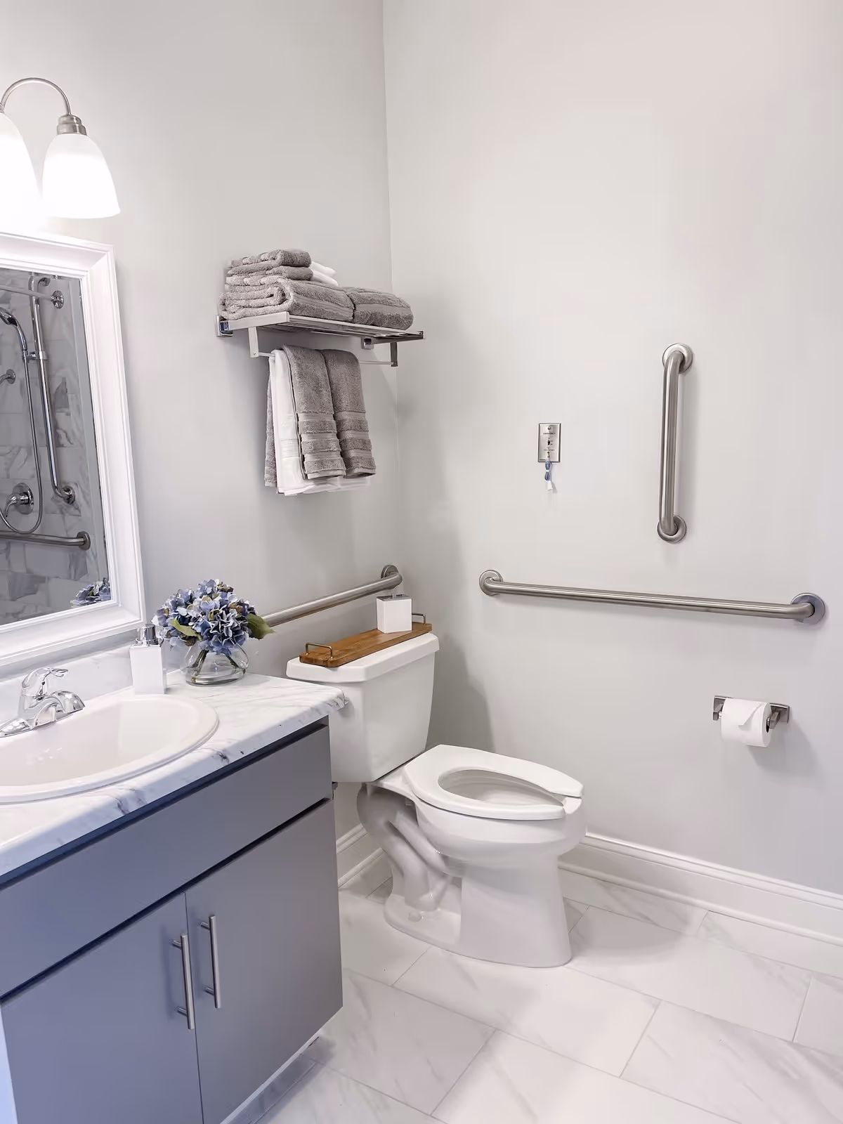 A clean and modern bathroom with a white toilet, gray vanity with a white marble countertop, a sink, and a mirror with a light fixture above it. There are gray towels neatly folded and hanging on a metal rack above the toilet. The bathroom has white walls, white tiled floor, and metal grab bars installed near the toilet for accessibility. A small vase with purple flowers is placed on the countertop.