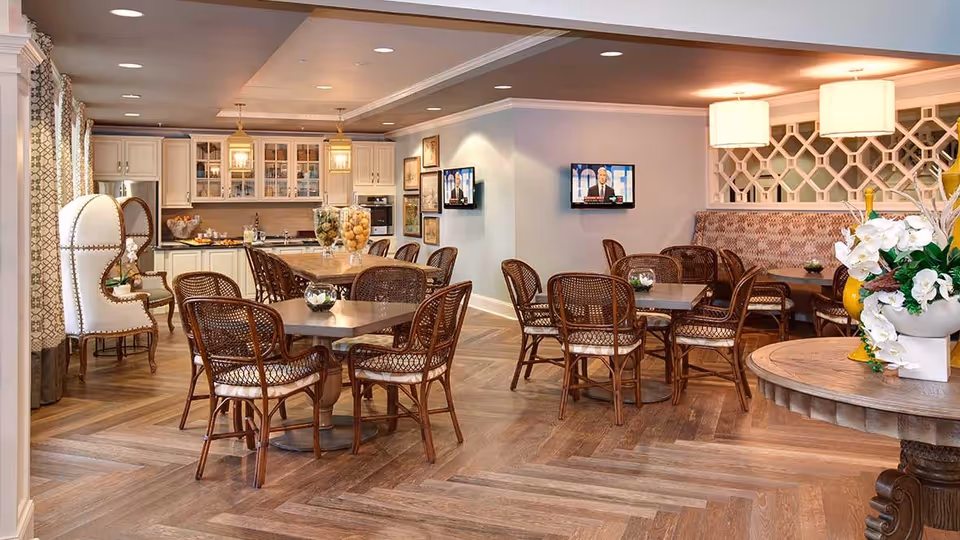 A cozy dining area in a senior living facility featuring multiple tables with wicker chairs, a kitchen area with white cabinets and glass doors, two wall-mounted televisions, decorative lighting fixtures, and floral arrangements on a round wooden table.