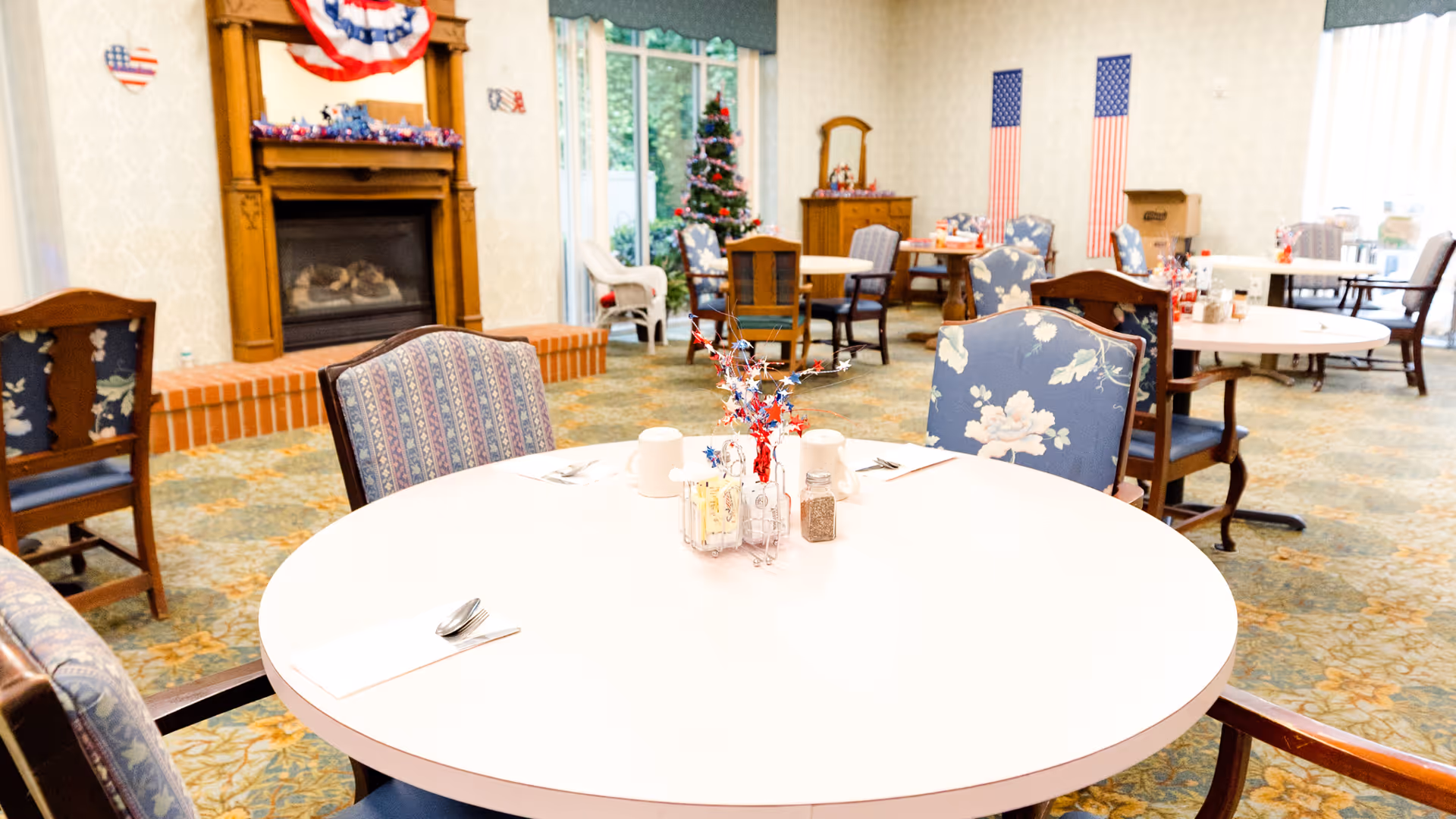 A bright dining room with round tables and chairs arranged neatly. The tables have simple place settings with napkins, spoons, and small condiment containers. In the background, there is a fireplace decorated with patriotic bunting and a Christmas tree near large windows. The room has a patterned carpet and light-colored walls with two American flags hanging on the far wall.