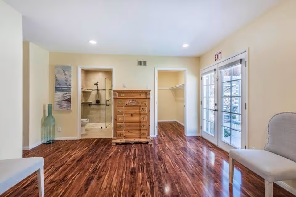 Interior room with polished wooden floor, light yellow walls, and recessed ceiling lights. There is a wooden chest of drawers centered between two doorways; the left doorway leads to a bathroom with a glass shower door, and the right doorway leads to a walk-in closet. To the right, there are double glass doors with an exit sign above, and two upholstered chairs are partially visible on the left and right edges of the image. A tall decorative glass vase and a wall painting are on the left side of the room.