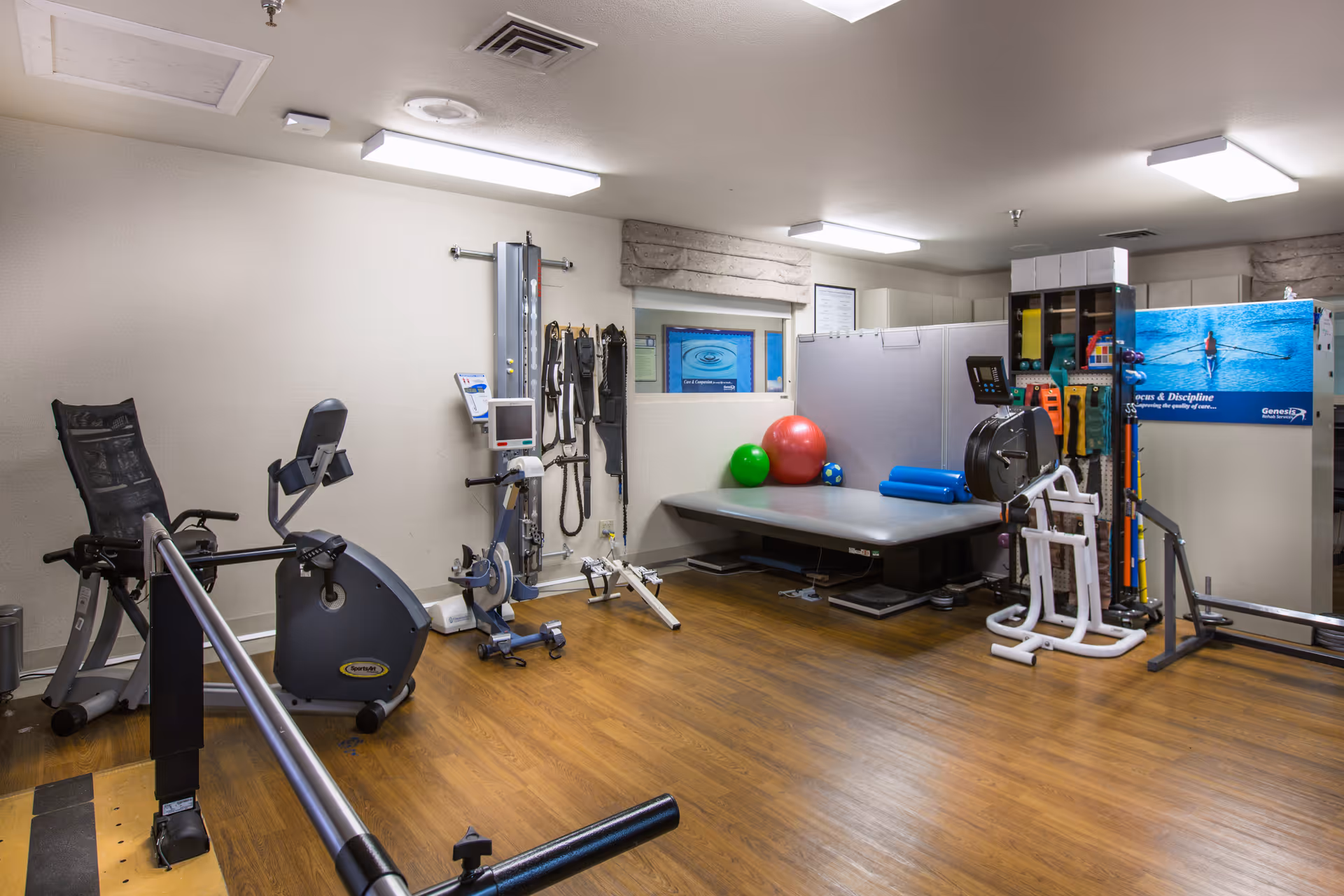 A rehabilitation therapy room with exercise equipment including a stationary bike, parallel bars, therapy balls, resistance bands, and a padded therapy table. The room has wood flooring, white walls, and bright overhead lighting.