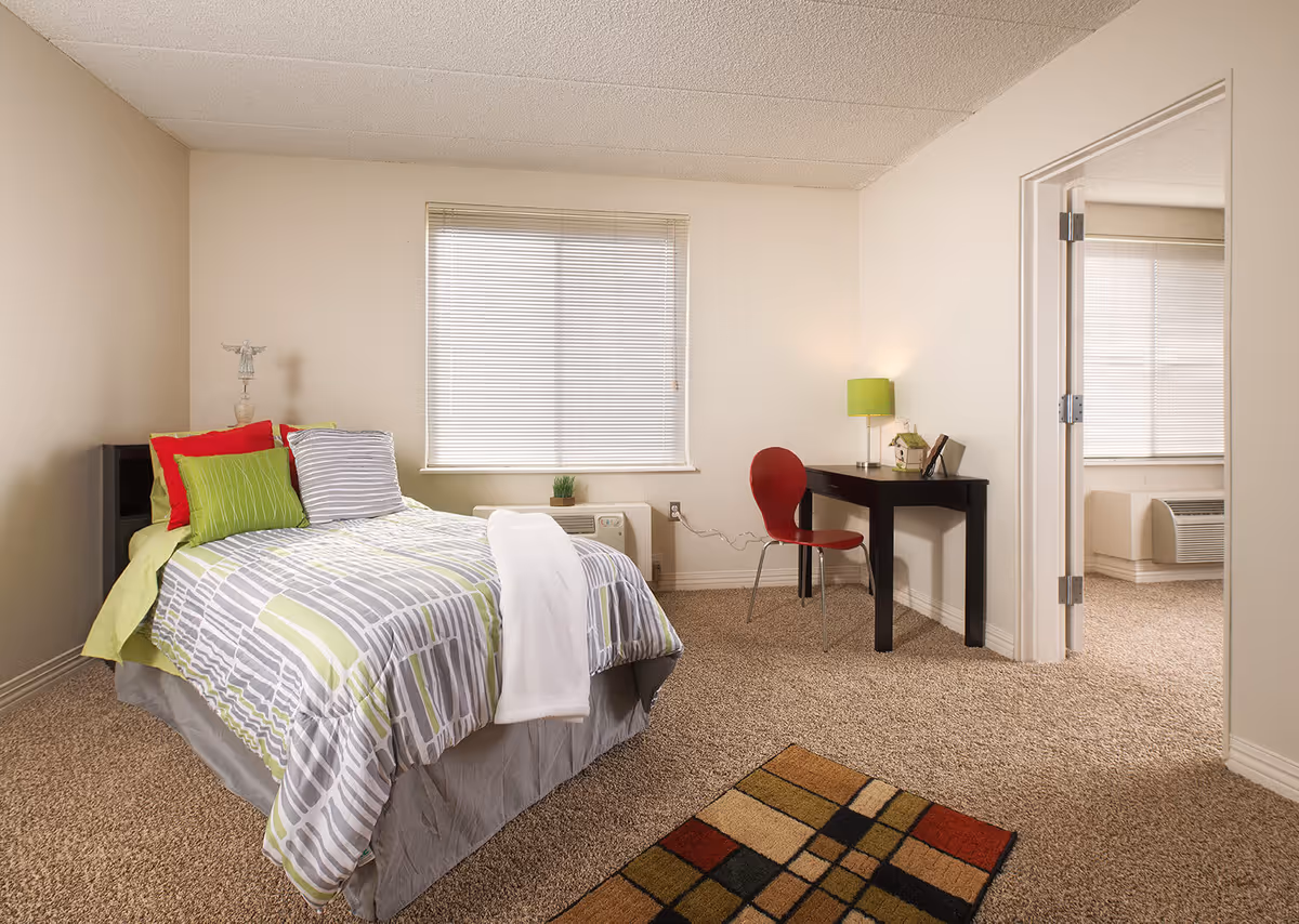 A cozy bedroom with a bed featuring green, gray, and red pillows and a patterned bedspread. There is a small black desk with a green lamp and a red chair next to it. A window with blinds is centered on the wall behind the bed, and an open doorway leads to another room with a similar window and air conditioning unit. The floor is carpeted with a small colorful rug near the bed.
