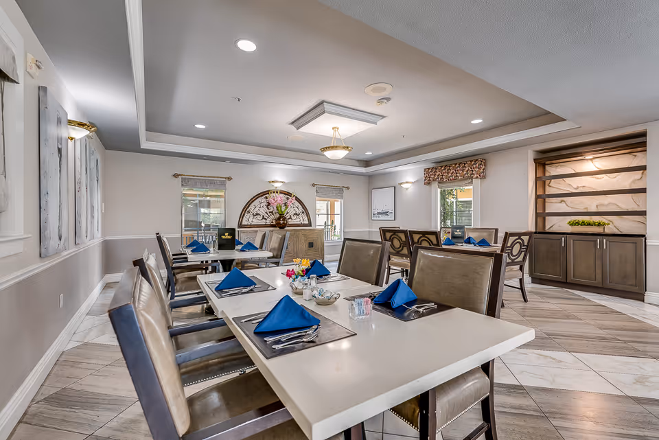 A bright and spacious dining room in a senior living facility with several tables set with blue napkins, silverware, and small flower arrangements. The room features beige walls, large windows with valances, decorative wall art, and a ceiling with recessed lighting and a central chandelier. There is a cabinet with a decorative backdrop on one side of the room.