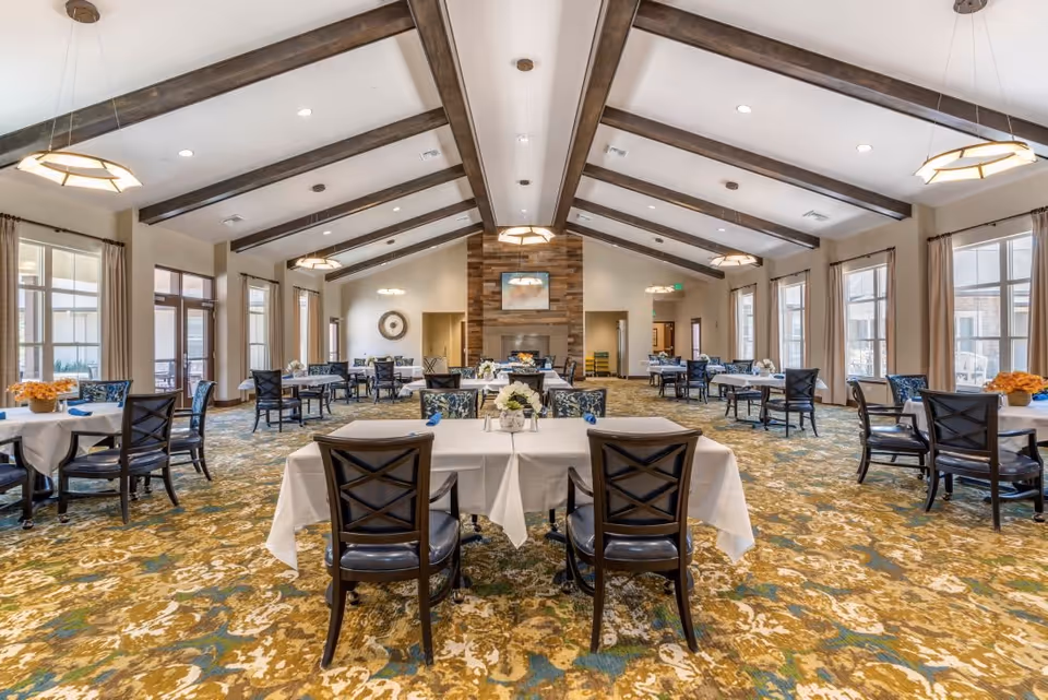 A spacious dining room with multiple tables covered in white tablecloths and surrounded by dark wooden chairs. The room features a high ceiling with exposed wooden beams and modern pendant lights. Large windows with beige curtains line the walls, allowing natural light to fill the space. A stone fireplace is centered on the far wall, above which hangs a framed picture. The carpet has a patterned design in shades of green, gold, and blue.