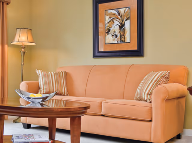 A cozy living room area with a peach-colored sofa adorned with two striped pillows. A wooden coffee table with a decorative bowl containing lemons is in front of the sofa. A floor lamp with a beige shade stands to the left, and a framed artwork featuring a palm tree hangs on the light green wall behind the sofa.