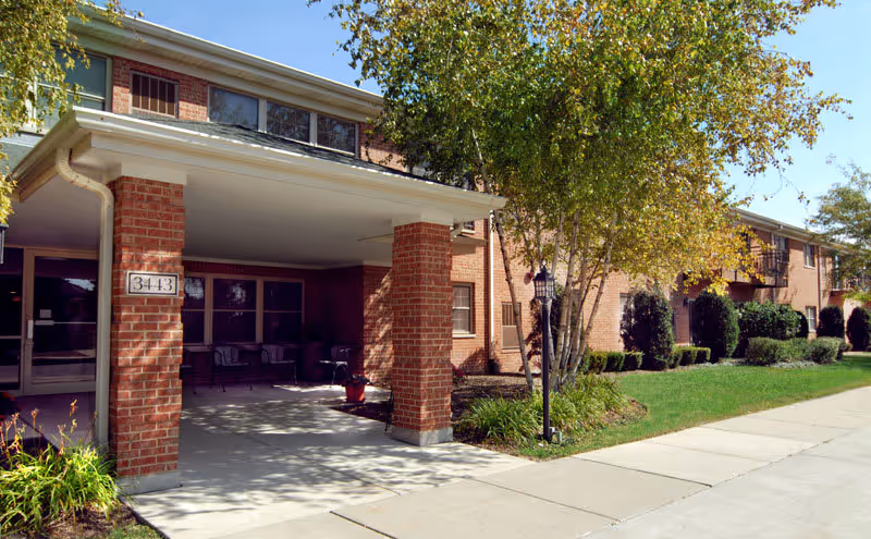 Exterior view of a brick senior living facility with a covered entrance supported by brick columns, a sidewalk, green lawn, trees, and shrubs under a clear blue sky.