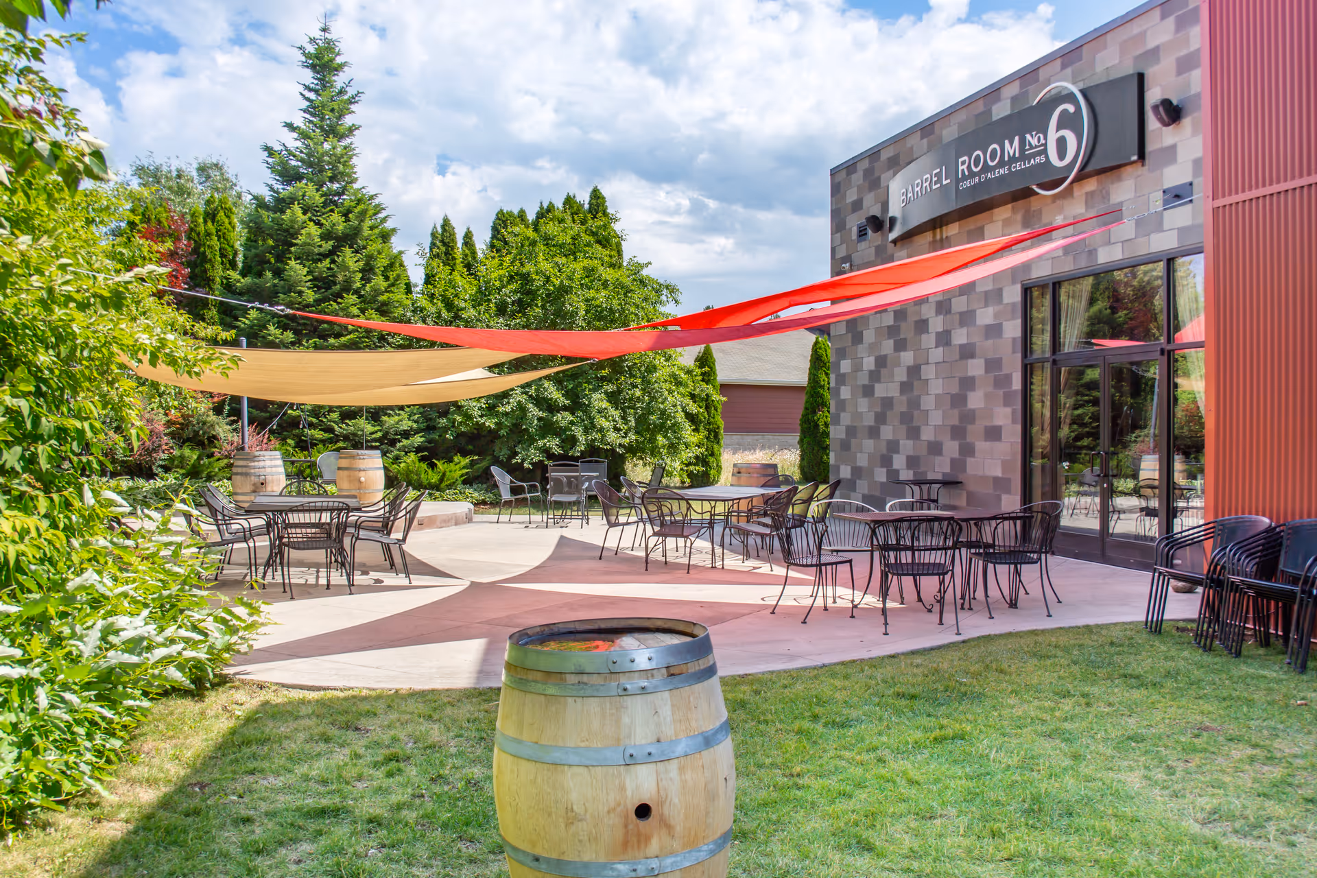 Outdoor patio area with metal tables and chairs under red and beige shade sails, surrounded by green trees and bushes. A wooden barrel is in the foreground and a building with a sign reading 'Barrel Room No 6 Coeur d'Alene Cellars' is visible in the background.