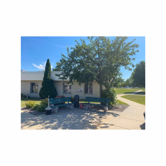Outdoor seating area with two green benches arranged in an L shape under a tree, next to a building with beige brick walls and windows. There is a trash can and a small planter between the benches. A paved walkway curves around the area, and the sky is clear and blue.