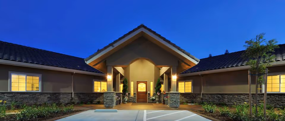 Front entrance of a single-story senior living building at dusk with lit windows, covered porch, driveway, and landscaping.