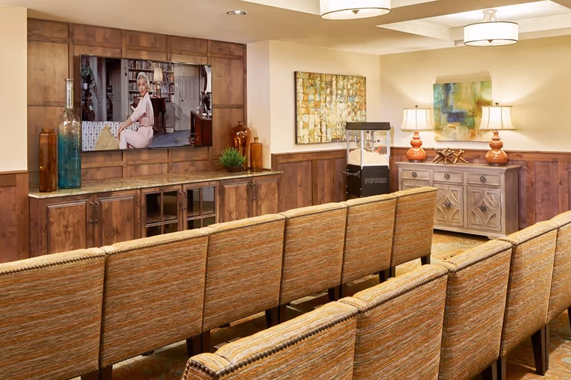 Rows of upholstered chairs face a wall-mounted TV in a cozy media/common room with wood cabinetry, artwork, lamps and a popcorn machine.