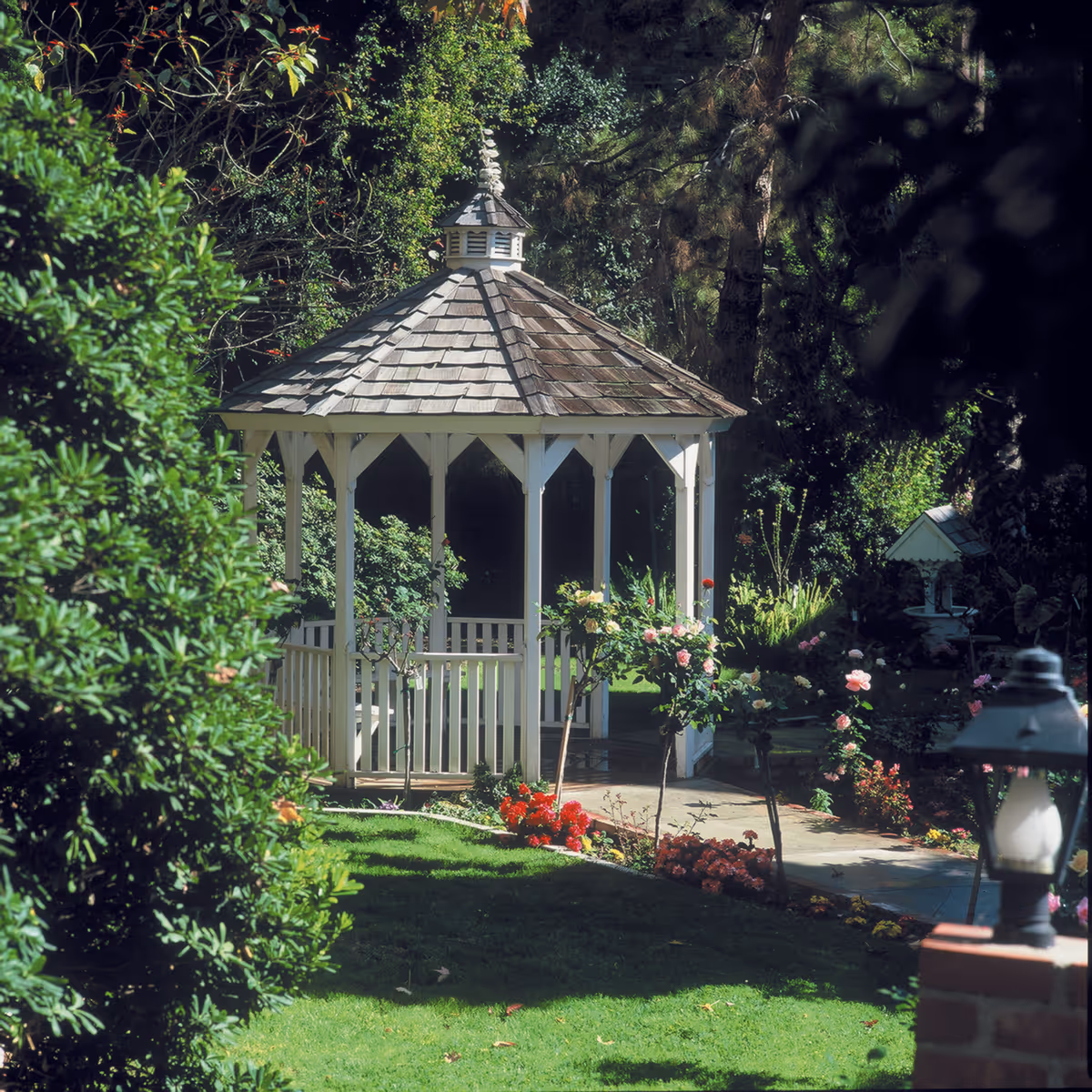 A white wooden gazebo with a shingled roof is situated in a lush garden surrounded by green bushes, trees, and colorful flowers. A paved pathway leads to the gazebo, and a decorative lamp post is visible in the foreground.