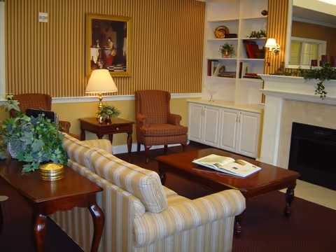 A cozy living room with a striped sofa, two upholstered armchairs, a wooden coffee table with an open book, a side table with a lamp and plant, built-in white shelves with decorative items, and a fireplace with a mantel decorated with greenery.