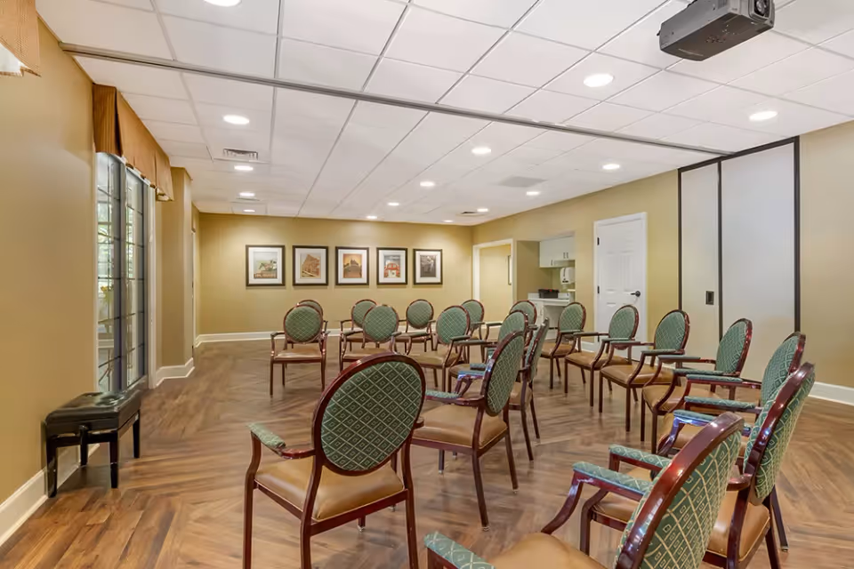 A well-lit meeting or activity room with multiple green cushioned chairs arranged in rows facing the front. The room has wood-patterned flooring, beige walls, and a white ceiling with recessed lighting. There are framed pictures on the far wall and a small counter area with cabinets and a door on the right side. Large windows with blinds are on the left side of the room.