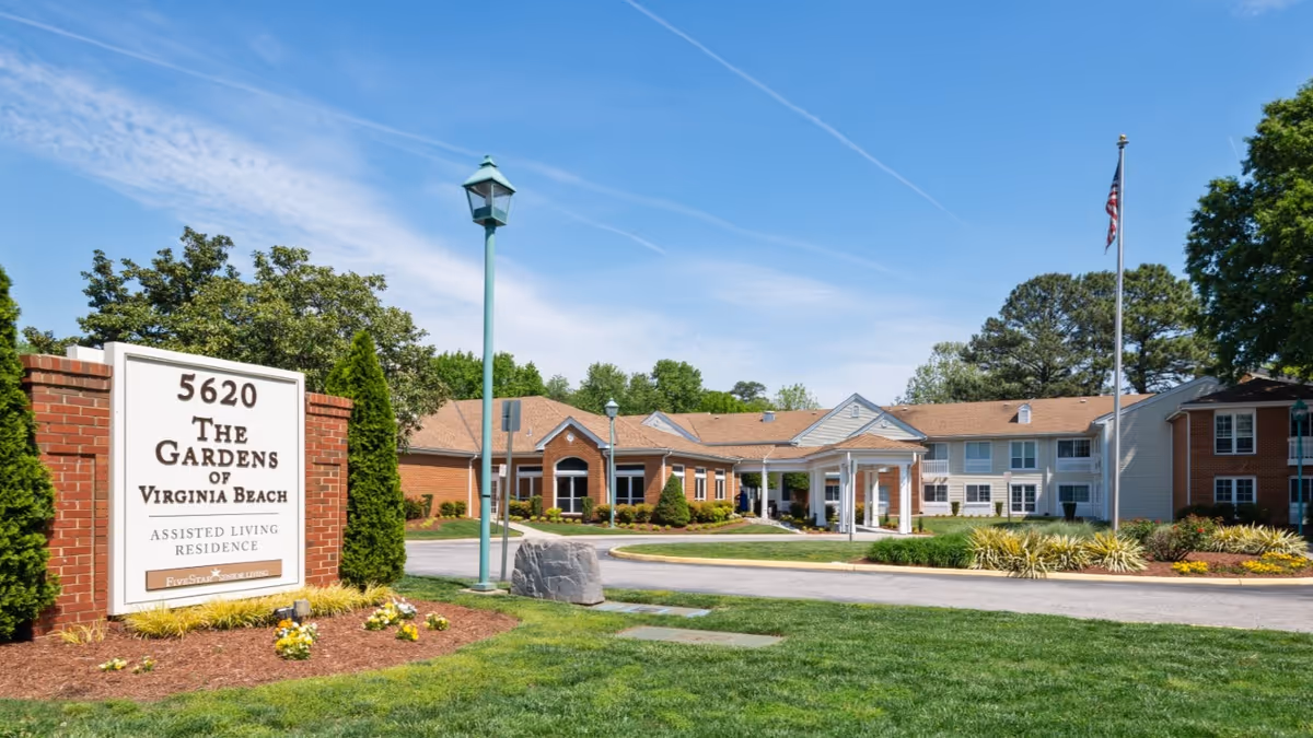 Exterior view of The Gardens of Virginia Beach assisted living residence showing the main building with a covered entrance, a flagpole with an American flag, a street lamp, and a large sign with the facility's name and address surrounded by landscaped greenery and flowers under a blue sky.