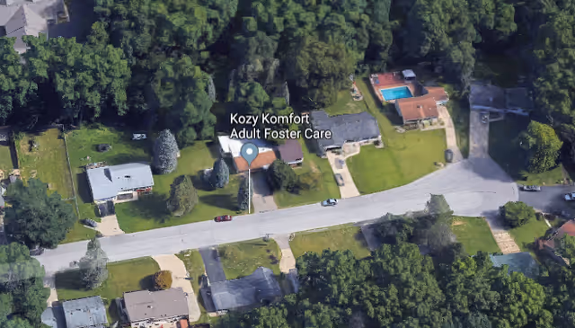Aerial view of a residential neighborhood showing Kozy Komfort Adult Foster Care facility with surrounding houses, trees, lawns, and a swimming pool in one backyard.