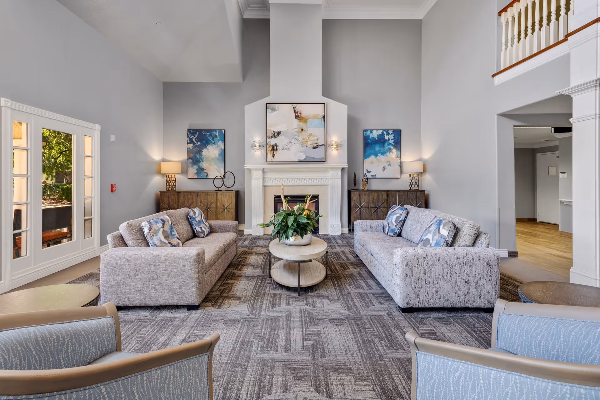 A spacious and well-lit living room with two gray sofas facing each other, a round coffee table with a plant in the center, two armchairs in the foreground, a fireplace with abstract artwork above it, and two matching cabinets with lamps and artwork on either side of the fireplace. Large windows on the left let in natural light.