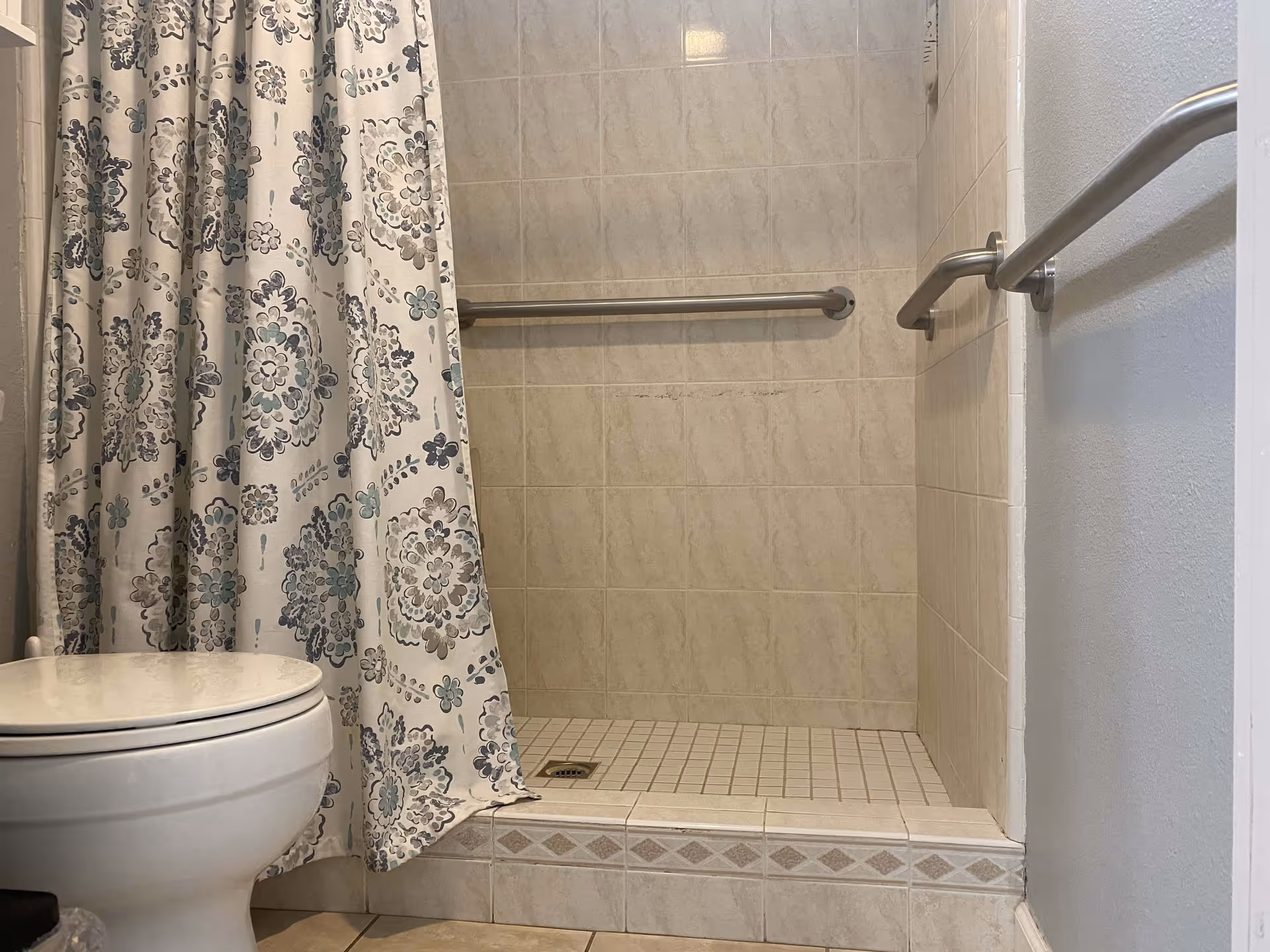 A bathroom with a white toilet on the left and a walk-in tiled shower with a floral patterned shower curtain partially drawn. The shower has beige tiles, a small tiled step, and two metal grab bars on the walls for support.