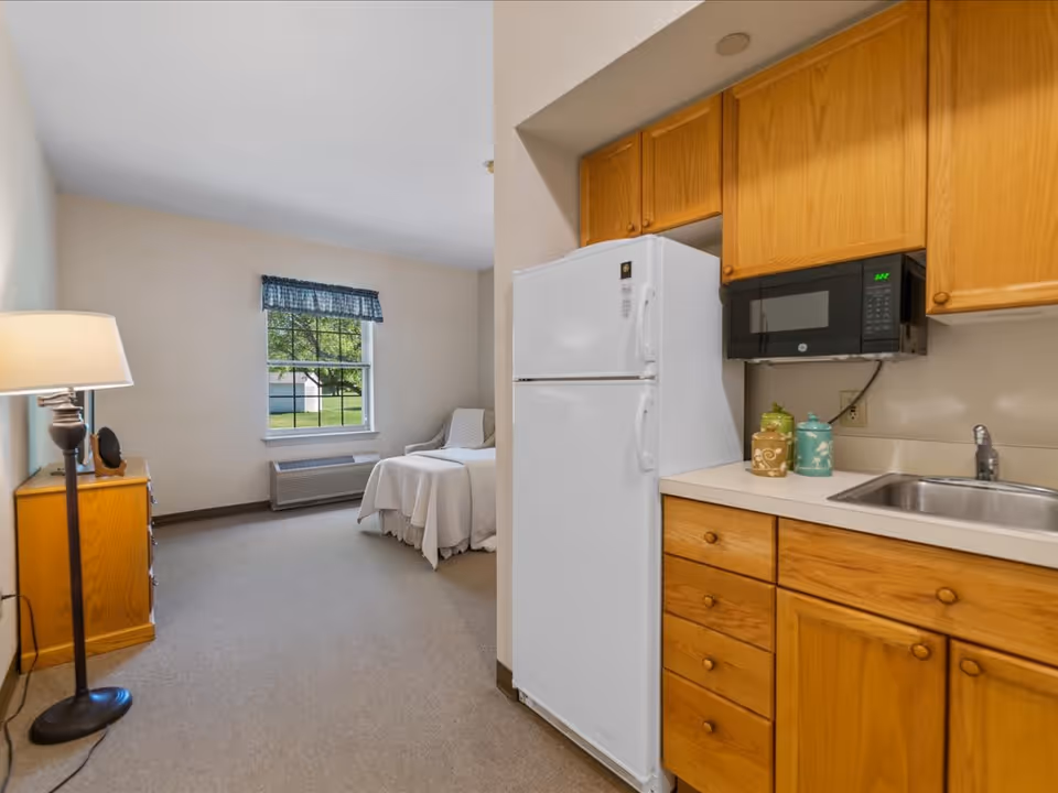 Interior view of a senior living facility room at Juniper Village at Mount Joy showing a small kitchen area with wooden cabinets, a white refrigerator, a microwave, and a sink. Adjacent to the kitchen is a bedroom area with a single bed covered in white bedding, a wooden dresser, a floor lamp, and a window with a blue valance overlooking green outdoor space.