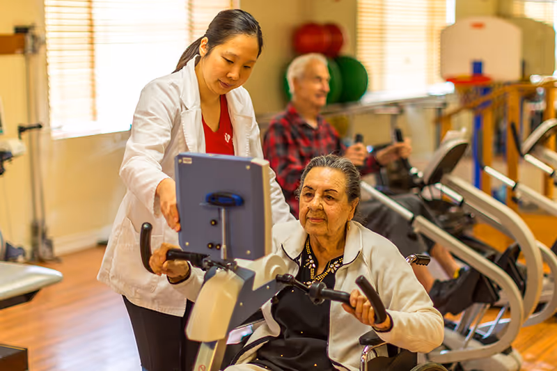 A healthcare professional assists an elderly woman using a stationary exercise bike in a rehabilitation center gym. In the background, an elderly man is also exercising on a similar machine. The room is well-lit with large windows and has exercise equipment and colorful therapy balls.