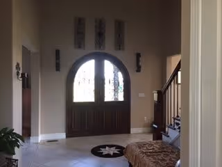 Interior view of an entryway in a residential or assisted living facility featuring a large double wooden door with decorative glass panels, beige walls, tiled floor, a round black rug with a white star pattern, a staircase with wooden railing on the right, and a cushioned bench or ottoman in the foreground.