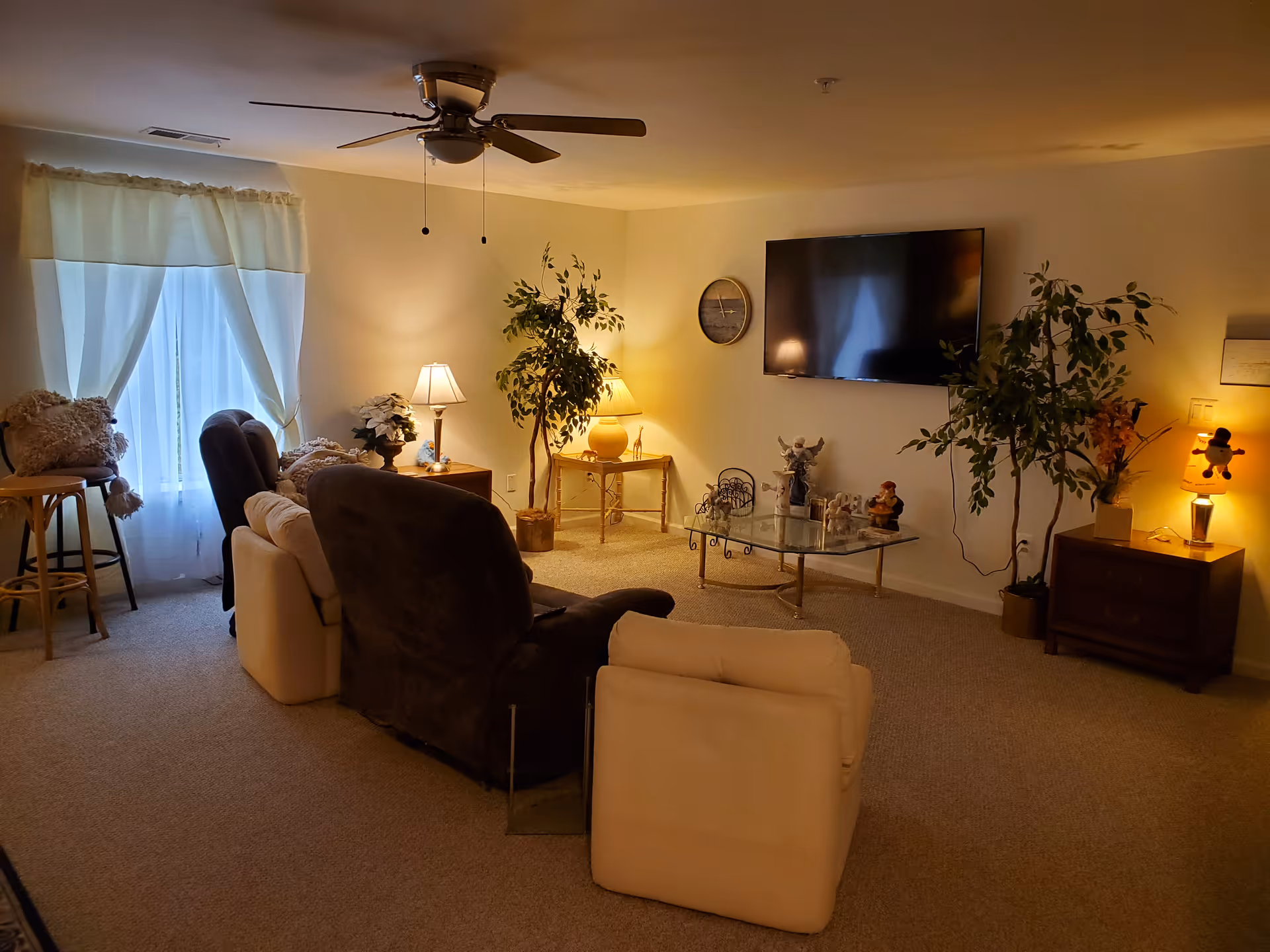 A cozy living room with beige carpet and cream-colored walls. The room features a ceiling fan with lights, a large flat-screen TV mounted on the wall, a glass coffee table with decorative items, two potted plants, and several lamps providing warm lighting. There are multiple comfortable chairs and a small table with a lamp and decorations near a window with sheer white curtains.