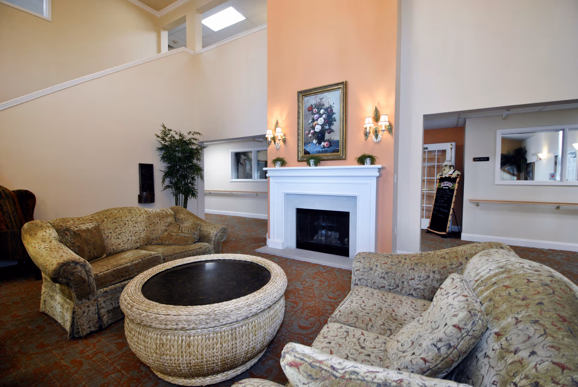 A comfortable common room with patterned sofas, a large round woven coffee table, and a white fireplace topped by a floral painting.