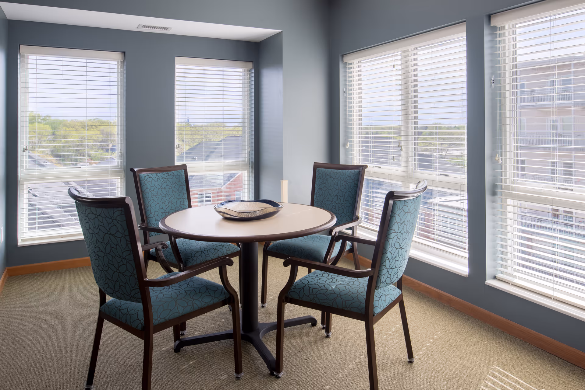 A small seating area with a round table and four upholstered chairs with wooden frames, positioned near large windows with white blinds. Outside the windows, rooftops and greenery are visible under a clear sky.