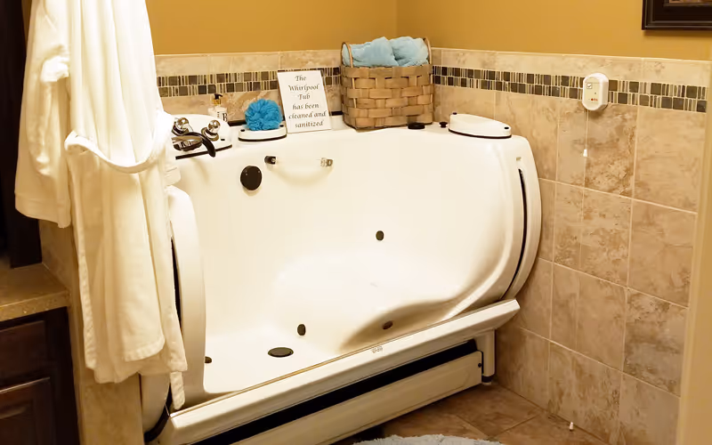 A clean and sanitized white walk-in whirlpool bathtub in a bathroom with beige tiled walls and floor. A white bathrobe hangs on the left side, and a basket with blue towels is placed on the tub's edge. A small sign on the tub reads 'The Whirlpool Tub has been cleaned and sanitized.'