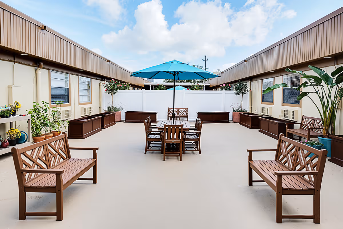 Outdoor courtyard area with wooden benches and a table with chairs under a blue umbrella, surrounded by potted plants and a white fence, under a partly cloudy sky.