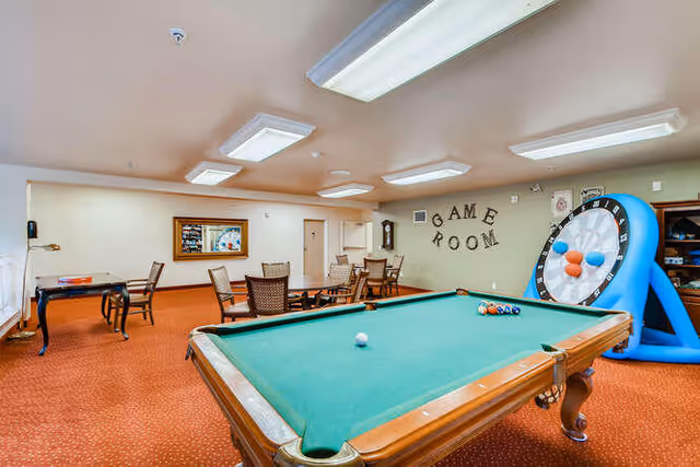 A game room with a pool table in the foreground, several chairs and tables in the background, a large inflatable dartboard game on the right, and a wall decoration spelling 'GAME ROOM'. The room has beige walls, a red carpet, and ceiling lights.