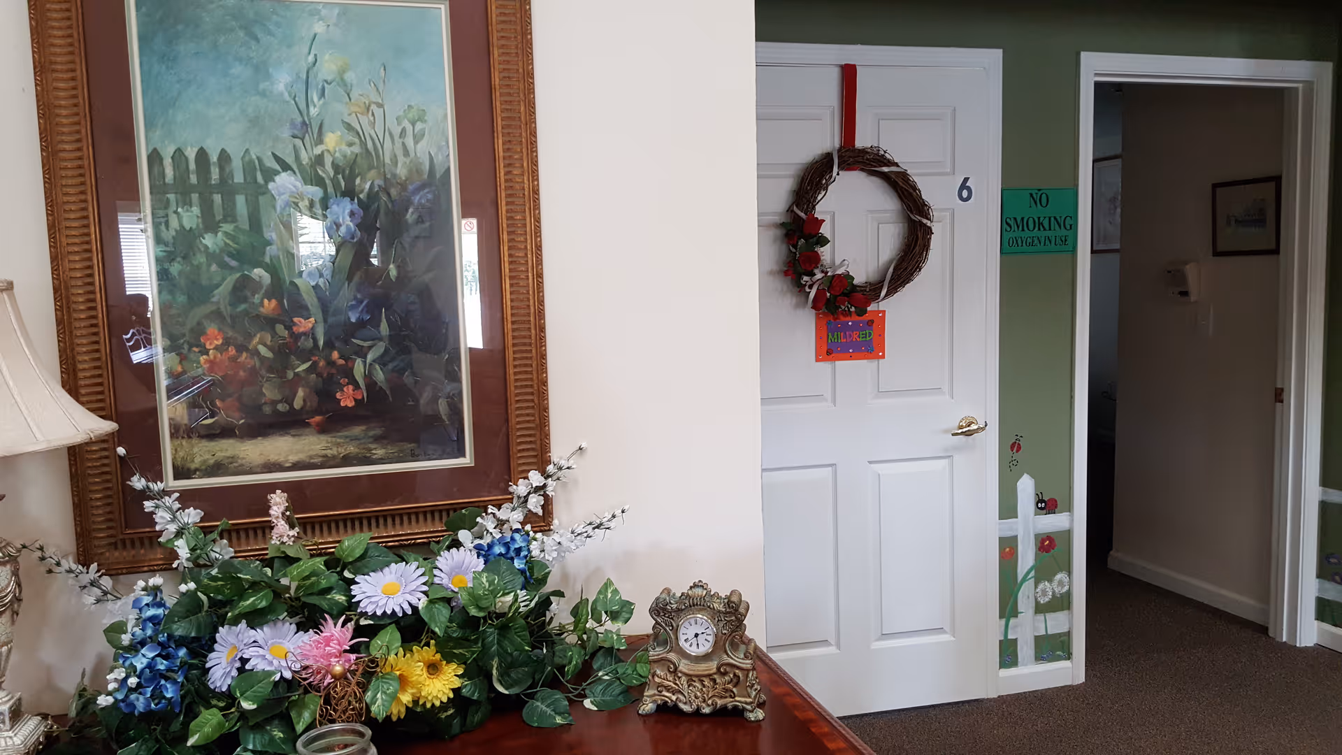 Interior view of a hallway in a senior living facility. A white door with the number 6 has a decorative wreath and a colorful sign that says 'Welcome'. To the left, there is a wooden table with a floral arrangement and an ornate clock. Above the table hangs a framed painting of flowers and a picket fence. The walls are painted green with a white chair rail and floral decorations near the base. A green sign on the wall reads 'No Smoking Oxygen in Use'.