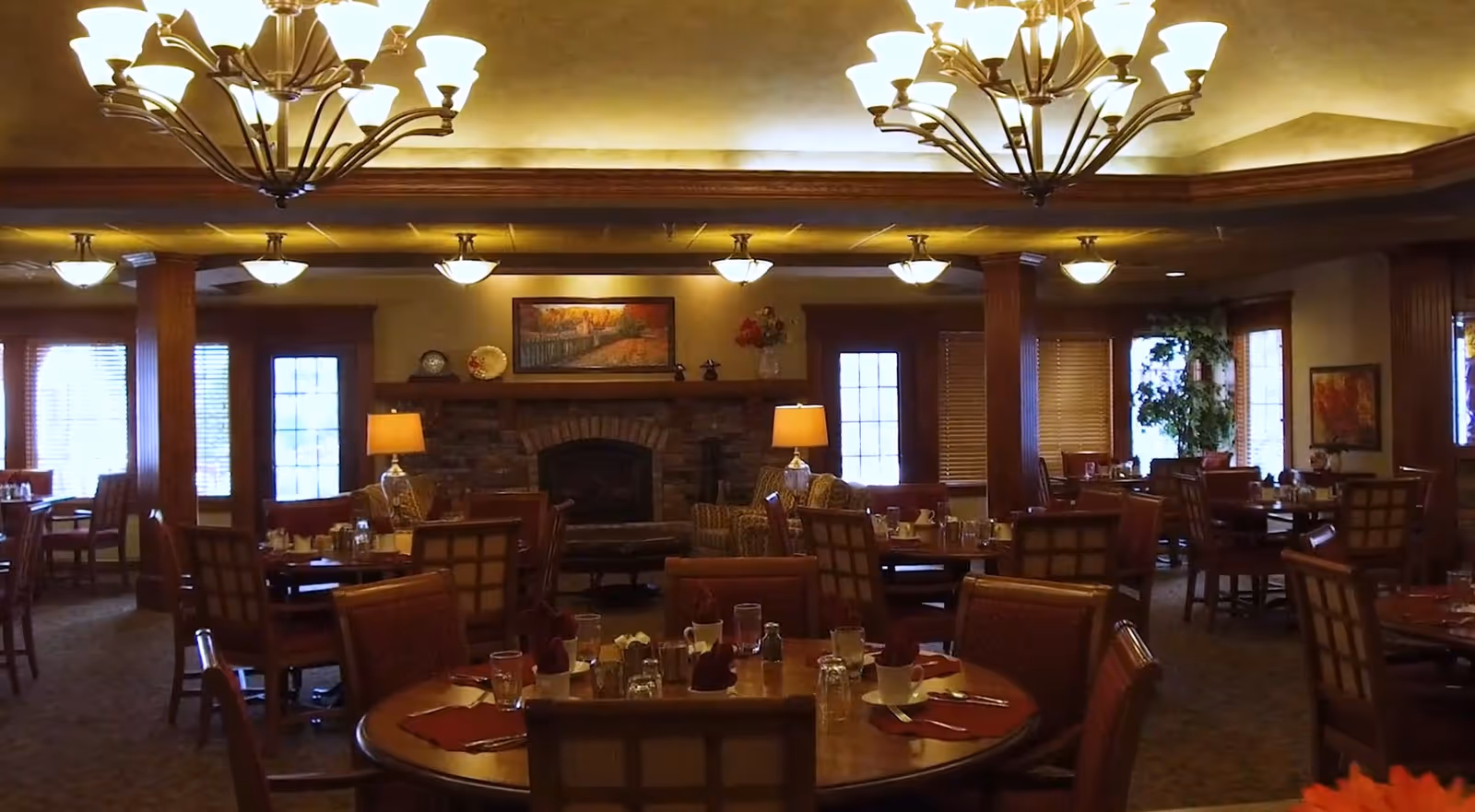 Warm, wood-accented dining room with round tables set for meals, chandeliers, and a central fireplace.