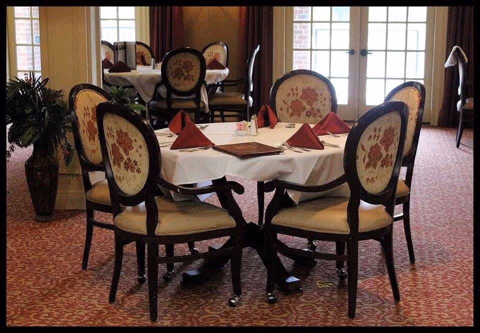 A dining area with a round table covered with a white tablecloth, set with red folded napkins, silverware, and a menu. The chairs around the table have floral upholstery. In the background, there are more tables and chairs, a large window with glass doors, and a potted plant on the left side.