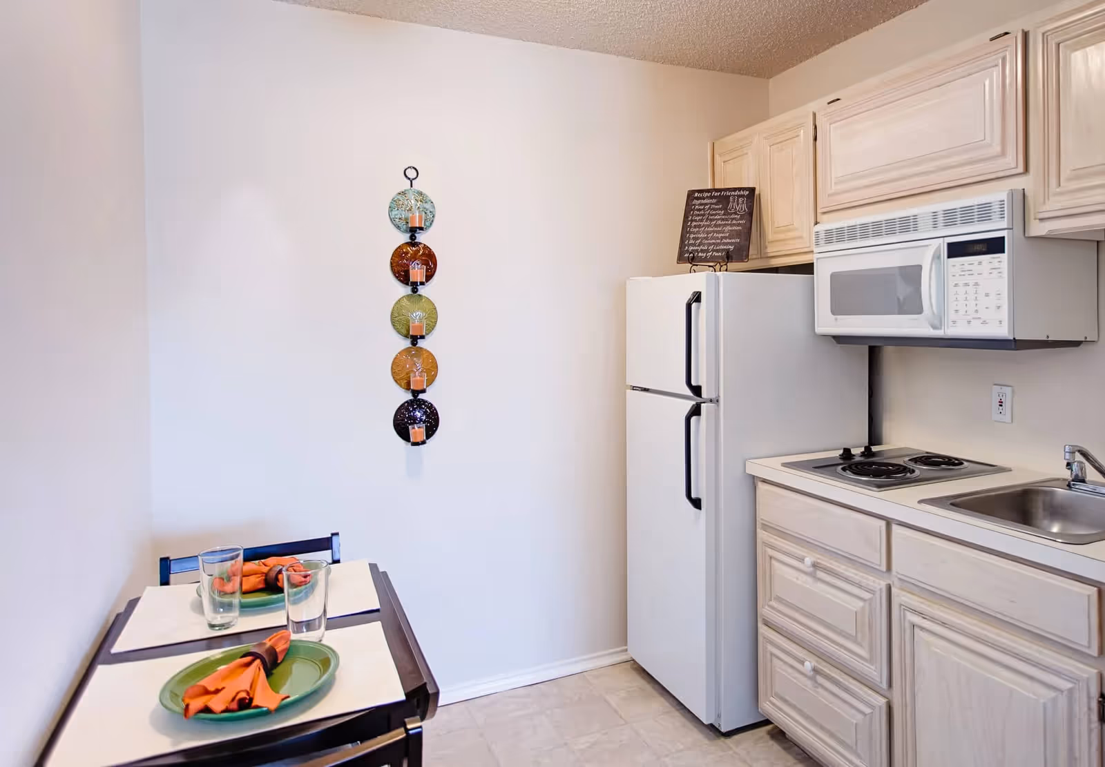 Small kitchen area with light wood cabinets, a white refrigerator, a white microwave above a two-burner electric stove, and a stainless steel sink. To the left, there is a small dining table set for two with green plates, orange napkins, and clear drinking glasses. A decorative wall hanging with five colorful circular plates holding small candles is mounted on the wall.