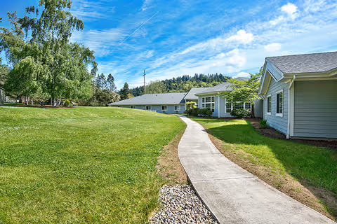 A paved walkway curves through a well-maintained grassy lawn leading to single-story buildings with gray siding and white trim under a blue sky with scattered clouds. Trees and hills are visible in the background.