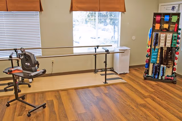 A rehabilitation or physical therapy room with parallel bars for walking exercises, a stationary exercise bike, and a rack holding various exercise and therapy equipment such as resistance bands and weights. The room has wooden flooring, two windows with blinds and brown valances, and light-colored walls.