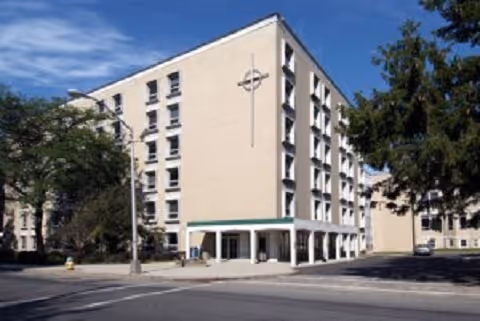 Front view of a multi-story beige building with a covered entrance, a cross emblem on the façade, trees, and a street in front.