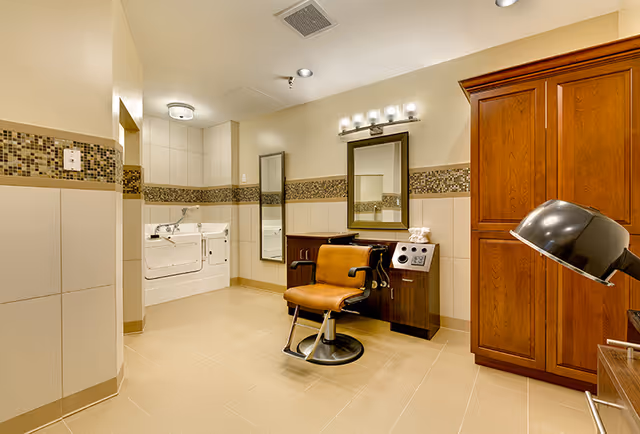 Interior view of a senior living facility room with a brown salon chair in front of a mirror and vanity, a large wooden cabinet, a hair dryer hood, and a walk-in bathtub with grab bars. The walls have beige tiles with a decorative mosaic tile border.