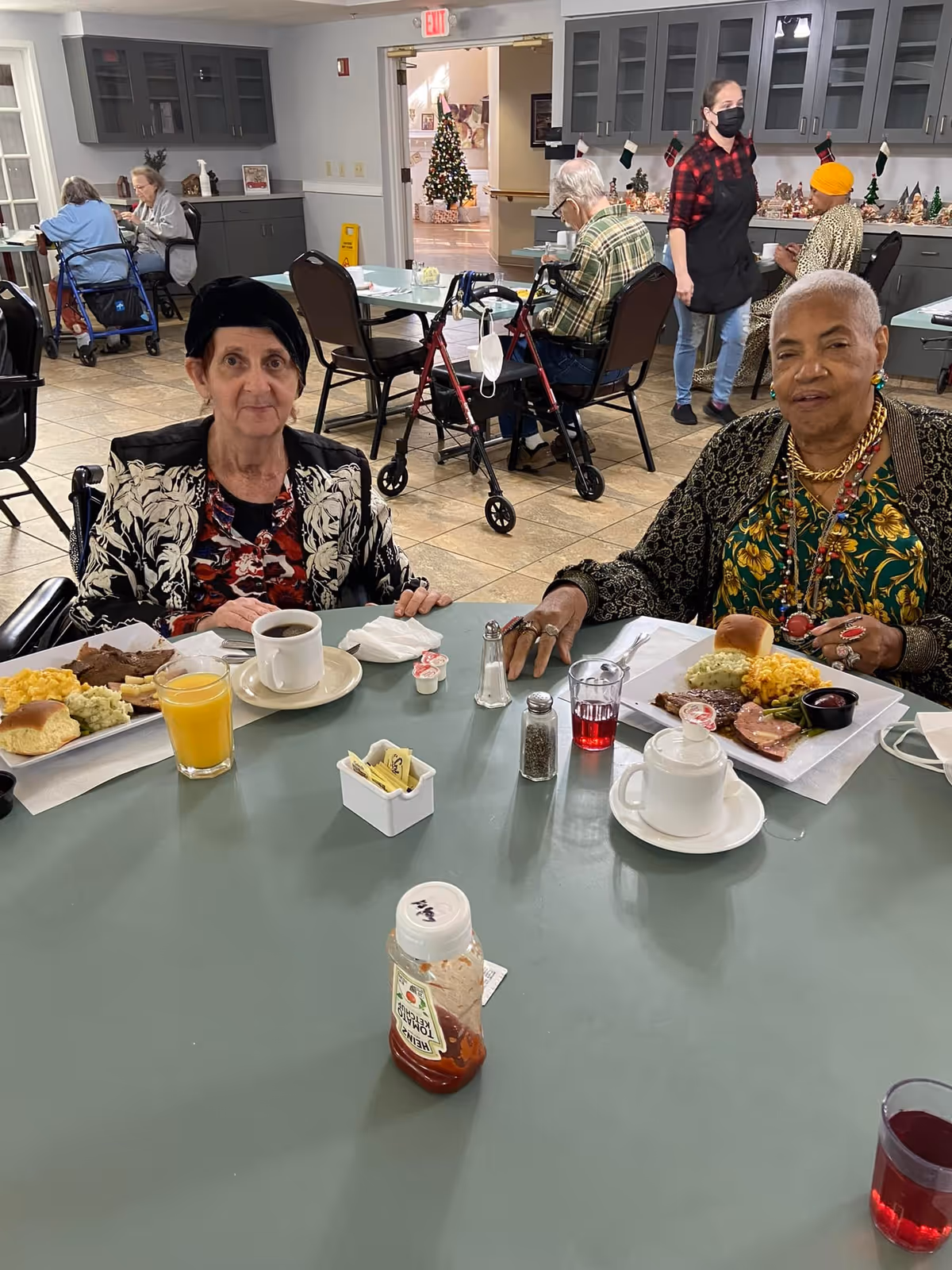 Two elderly women sitting at a dining table in a senior living facility, enjoying a meal with plates of food, coffee, and juice. In the background, other seniors are seated at tables, and a staff member wearing a mask and apron is standing. The room has gray cabinets and a Christmas tree is visible through an open doorway.