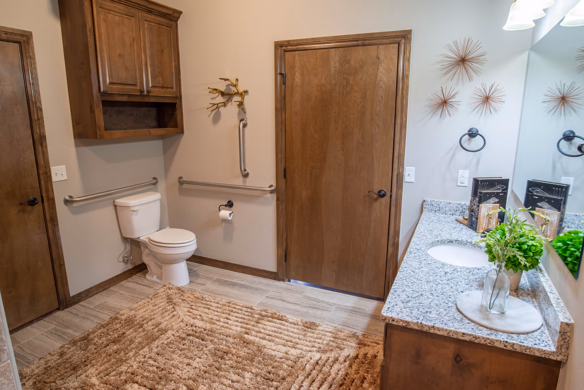 A clean and spacious bathroom with a white toilet, wooden cabinets, a granite countertop with a sink, a large mirror, and decorative wall art. The floor has light-colored tiles and a textured beige rug. There are safety grab bars installed near the toilet and on the walls.