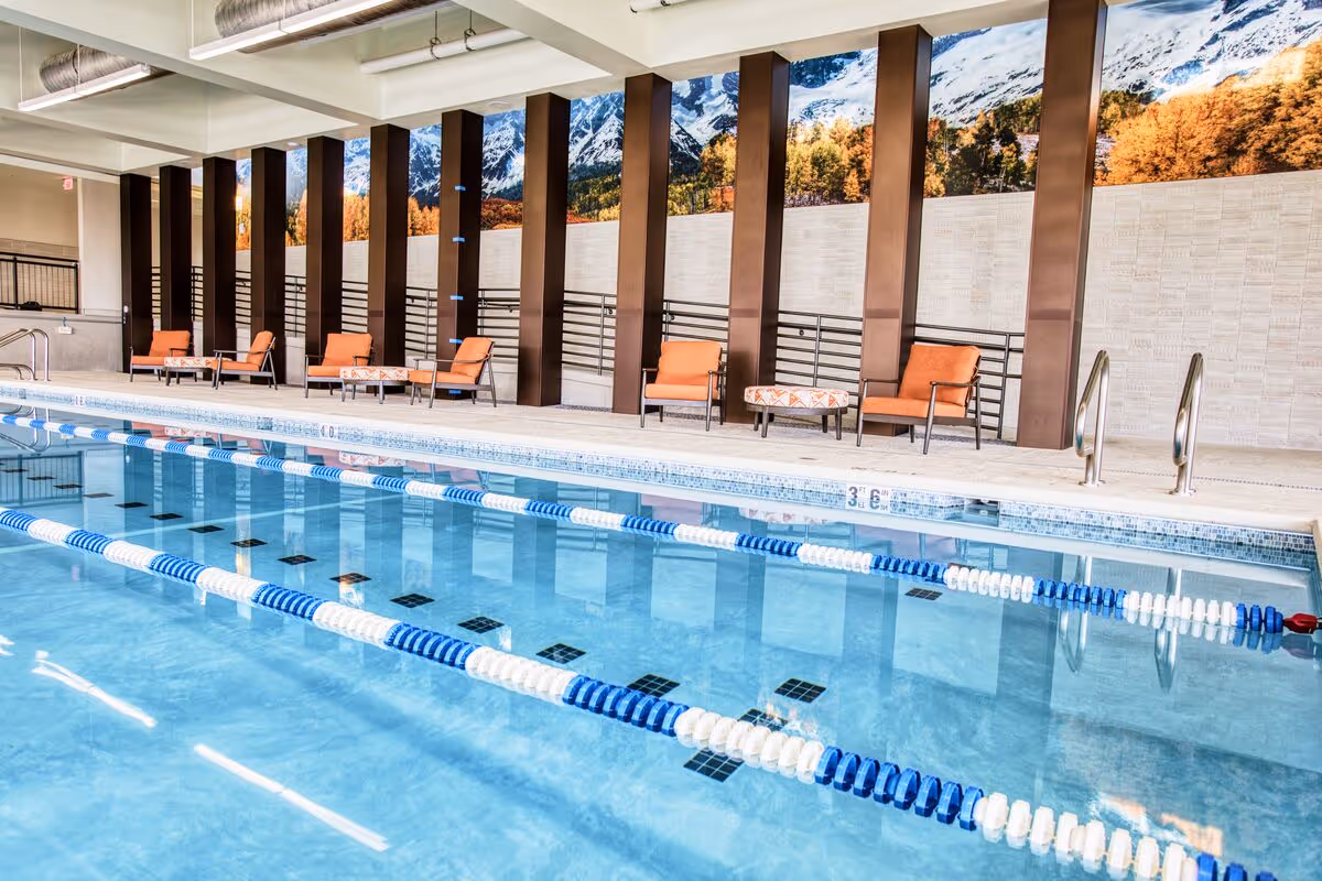 Indoor swimming pool with blue and white lane dividers, several orange cushioned lounge chairs and small tables along the poolside, and a mural of snow-capped mountains and autumn trees on the wall behind the chairs.