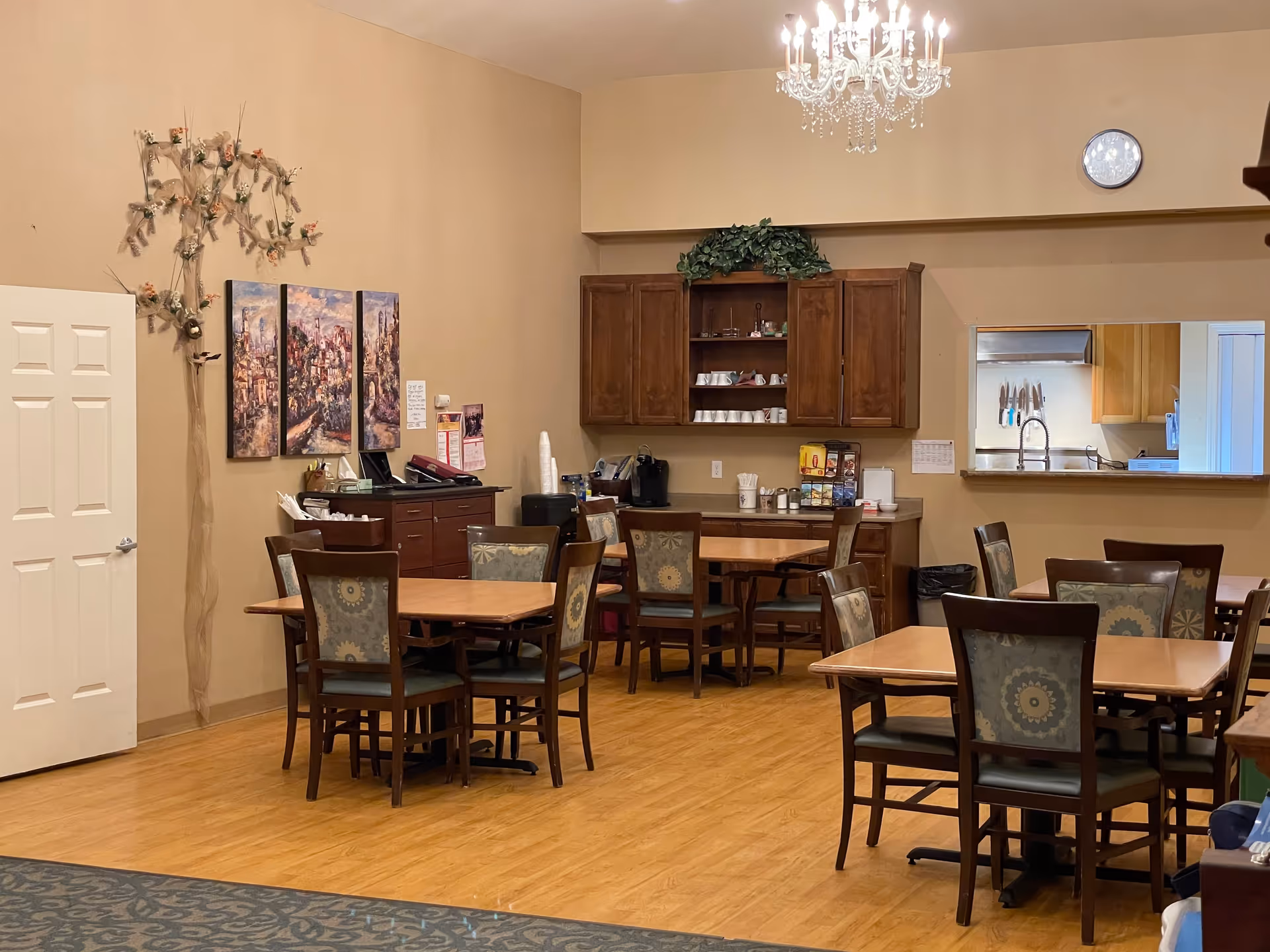 Dining room with several wooden tables and upholstered chairs, cabinets and a beverage station along a beige wall, and a chandelier overhead.